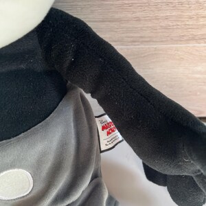 May include: A plush Mickey Mouse doll with a black body, gray shorts, and white gloves. The doll has a white face and a tag that reads "Disney Mickey Mouse". The doll's arm is extended.