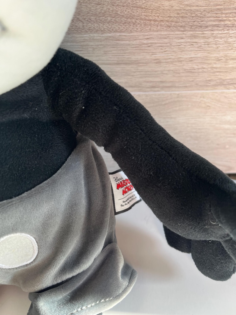 May include: A plush Mickey Mouse doll with a black body, gray shorts, and white gloves. The doll has a white face and a tag that reads "Disney Mickey Mouse". The doll's arm is extended.