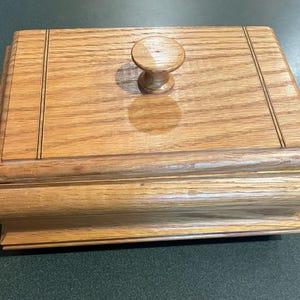 May include: A rectangular wooden box with a natural light brown grain finish and a smooth, polished surface. The lid features a centered, round wooden knob handle and two thin, parallel engraved lines running vertically across the top panel.