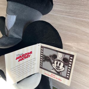 May include: A black, gray, and white Mickey Mouse plush toy with a tag featuring the text "Disney's Milestone Mickey" and "Steamboat Willie 1928". The tag also includes a black and white image of Mickey Mouse.