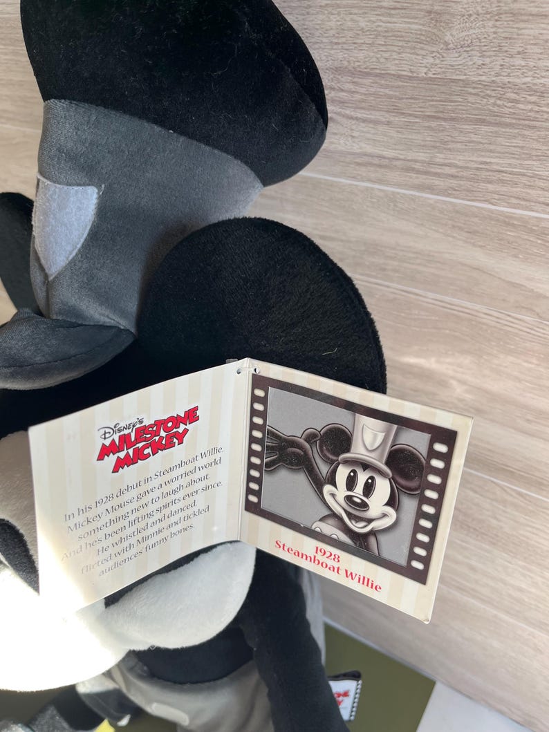 May include: A black, gray, and white Mickey Mouse plush toy with a tag featuring the text "Disney's Milestone Mickey" and "Steamboat Willie 1928". The tag also includes a black and white image of Mickey Mouse.