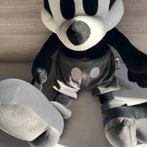 May include: A plush Mickey Mouse toy in black, white, and gray. The toy has large black eyes, a white face, and black ears. It wears gray shorts with two white buttons. The toy is a limited edition.