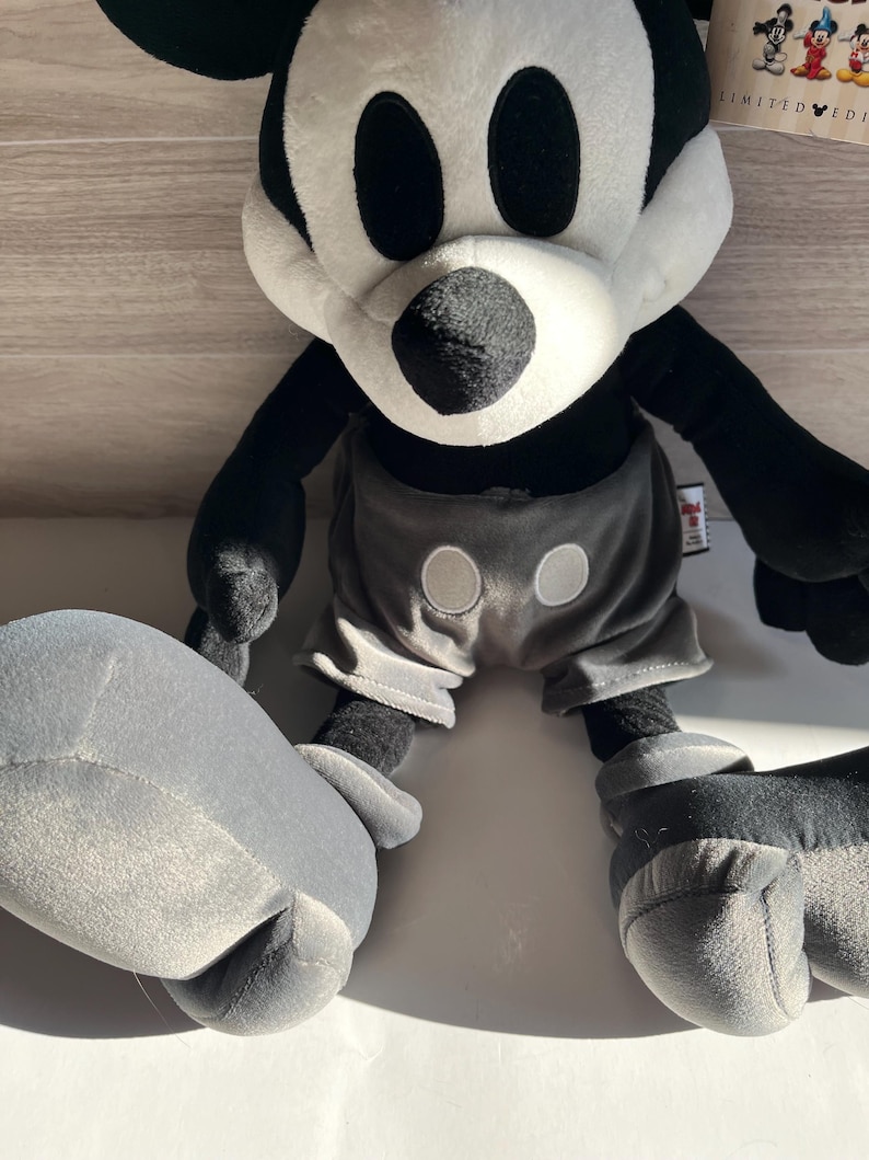 May include: A plush Mickey Mouse toy in black, white, and gray. The toy has large black eyes, a white face, and black ears. It wears gray shorts with two white buttons. The toy is a limited edition.