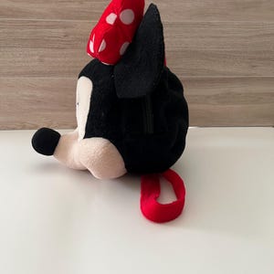 May include: A plush Minnie Mouse bag in black, white, and red. The bag features a red bow with white polka dots, black ears, and a red loop. The bag is lying on a white surface.