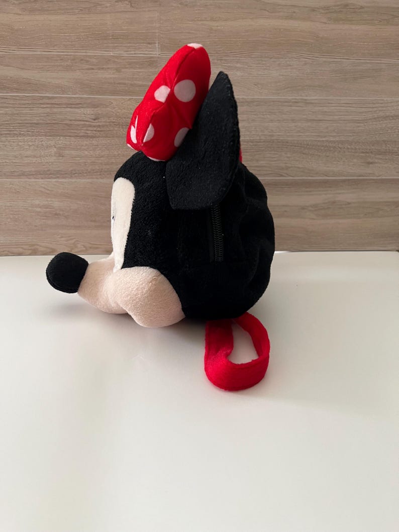 May include: A plush Minnie Mouse bag in black, white, and red. The bag features a red bow with white polka dots, black ears, and a red loop. The bag is lying on a white surface.