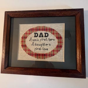 May include: A framed cross-stitch embroidery featuring a red, black, and tan plaid oval border on a light fabric background. The black text reads: DAD, A son's first hero, A daughter's first love. The piece is enclosed in a dark brown wooden frame.