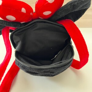 May include: Black plush backpack with red straps and a red bow with white polka dots. The backpack has a zippered closure and black ears. The straps are red.