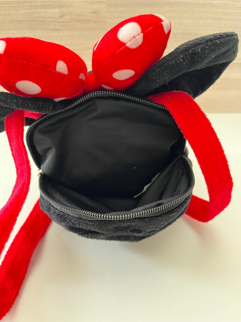 May include: Black plush backpack with red straps and a red bow with white polka dots. The backpack has a zippered closure and black ears. The straps are red.
