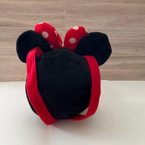 May include: A black plush bag with a red strap and a red bow with white polka dots. The bag has large black ears and a zipper closure. The bag is designed to resemble a cartoon character.