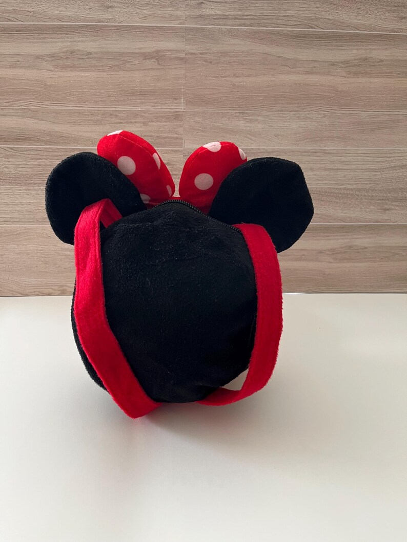 May include: A black plush bag with a red strap and a red bow with white polka dots. The bag has large black ears and a zipper closure. The bag is designed to resemble a cartoon character.