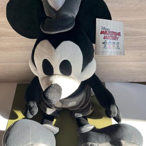 May include: A plush Mickey Mouse toy in black, white, and gray, with a hat and shorts. The toy has large ears and a tag that reads "Disney's Milestone Mickey Limited Edition."