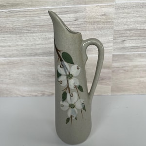 May include: A tall, slender ceramic pitcher with a matte, speckled light gray finish. The pitcher features a hand-painted design of white dogwood flowers with green leaves and brown stems on the front, a curved handle, and a pointed pouring spout.