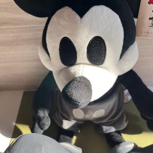 May include: A plush Mickey Mouse toy in black, white, and gray. The toy has large black eyes, a black nose, and a gray hat. The toy is wearing gray shorts and white gloves. The toy is a limited edition.