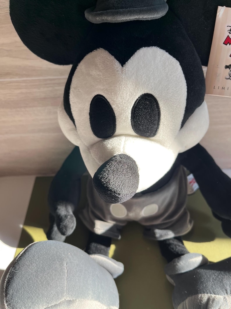 May include: A plush Mickey Mouse toy in black, white, and gray. The toy has large black eyes, a black nose, and a gray hat. The toy is wearing gray shorts and white gloves. The toy is a limited edition.