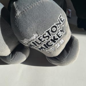 May include: Close-up of a gray plush toy's foot with the embroidered text "MILESTONE MICKEY 1928". The toy is a soft, stuffed figure, likely a collectible item.