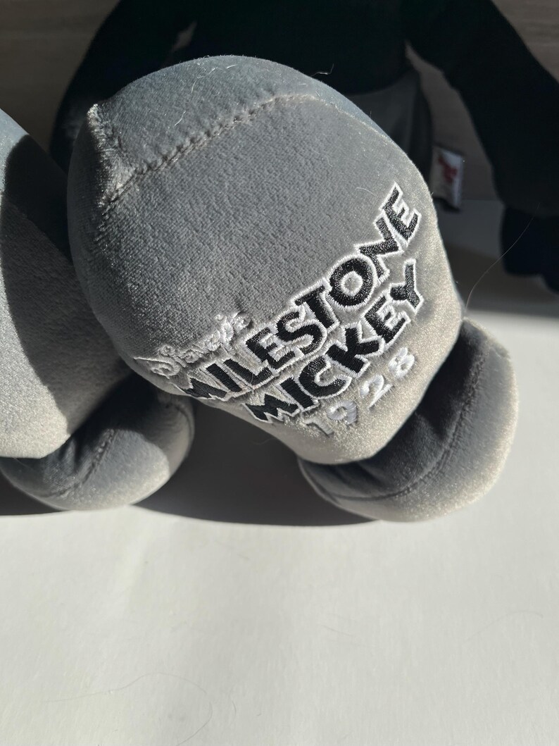 May include: Close-up of a gray plush toy's foot with the embroidered text "MILESTONE MICKEY 1928". The toy is a soft, stuffed figure, likely a collectible item.