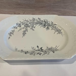 May include: Oval white serving platter with a floral design in black and gray. The platter has a decorative embossed border. The floral design is centered on the platter and is a botanical illustration.