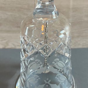 May include: Clear glass bell with etched bird and floral designs. The bell has a diamond-cut pattern and a silver-colored clapper. The base of the bell has the etched text "1987".