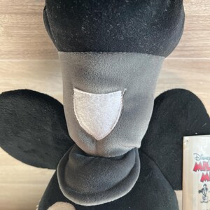May include: A plush Mickey Mouse doll with a black head and ears, a gray face and neck, and a white patch. The doll has black wings and a tag with the Disney logo. The doll is made of soft, velvety material.