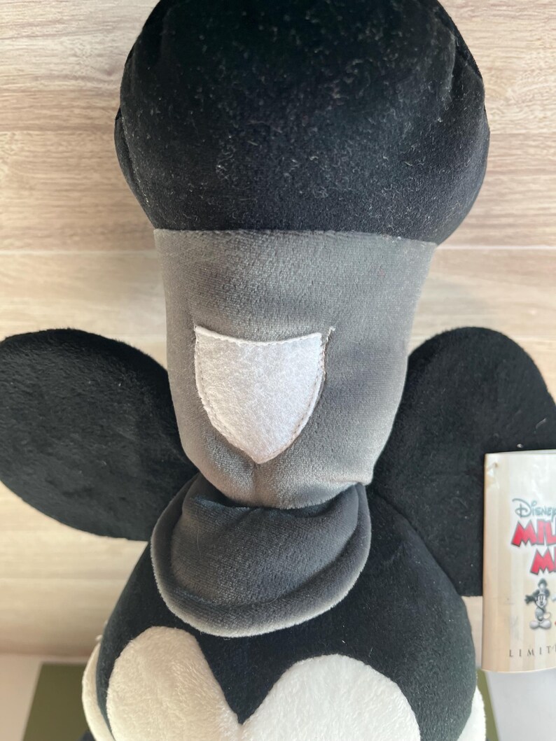 May include: A plush Mickey Mouse doll with a black head and ears, a gray face and neck, and a white patch. The doll has black wings and a tag with the Disney logo. The doll is made of soft, velvety material.