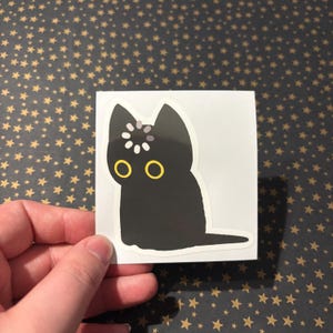 Può includere: Adesivo di un gatto nero con occhi gialli e un disegno bianco e grigio sulla testa. Il gatto è seduto con la coda distesa. L'adesivo è su sfondo bianco.