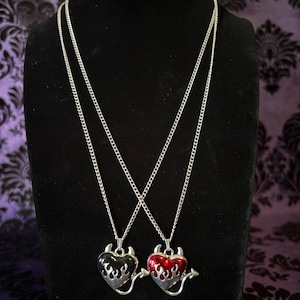 May include: Two silver necklaces with heart-shaped pendants. One pendant is black with silver flames, the other is red with silver flames. Each heart has devil horns and a tail.