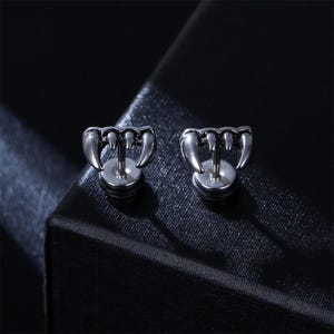 Puede incluir: Un par de pendientes de botón en tono plateado con un diseño de cuatro colmillos afilados y curvados dispuestos en fila. Los pendientes tienen un acabado metálico con detalles oscuros en relieve para resaltar la forma de los dientes.