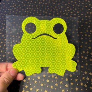 May include: A reflective frog sticker in bright neon green. The frog has large, round eyes and a curved mouth. The sticker is on a clear backing, and the background is a dark surface with gold stars.