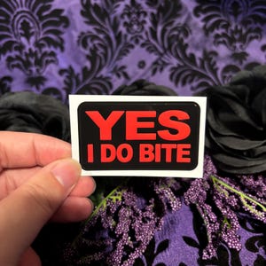 Peut inclure: Un autocollant rectangulaire avec un fond noir et une bordure blanche. Le texte 'YES I DO BITE' est imprimé en lettres capitales rouges vives et grasses au centre de l'autocollant, avec 'YES' sur la ligne du haut et 'I DO BITE' en dessous.