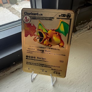Peut inclure: Une carte Pokémon Charizard dorée, évolution de stade 2, avec le texte "Charizard Lv.76". La carte présente un dragon orange flamboyant avec des ailes vertes. La carte est présentée sur un présentoir en plastique transparent.