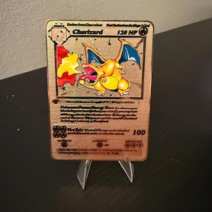 May include: A gold-colored Charizard Pokémon card, featuring a yellow dragon-like creature with blue wings and flames. The card has text including "Charizard" and "120 HP". It is displayed on a clear stand.