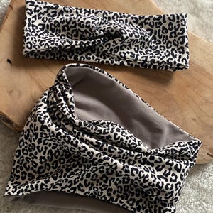 Könnte beinhalten: Ein Stirnband und ein Halstuch-Set. Beide Artikel weisen ein schwarz-weißes Leopardenmuster auf cremefarbenem Hintergrund auf. Das Halstuch hat ein graues Innenfutter. Die Artikel werden auf einer Holzoberfläche präsentiert.