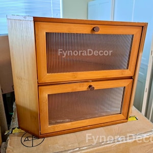 May include: A two-tiered wooden cabinet with clear, ribbed glass doors and round wooden knobs. The cabinet is made of light brown wood and features the text "FynoraDecor" on the glass doors.