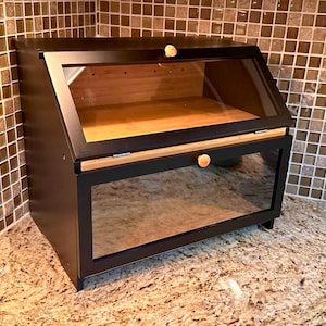 May include: A black bread box with a clear glass front and top. The box has a wooden interior and small wooden knobs. The bread box is sitting on a granite countertop.