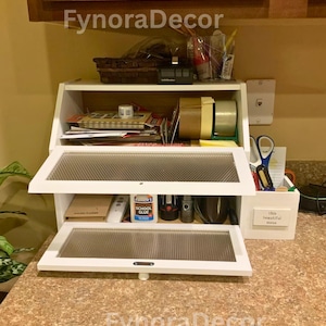 May include: A white, two-tiered desk organizer with glass-fronted compartments. The top shelf holds office supplies, while the bottom shelf contains glue and other items. The text "FynoraDecor" is visible.