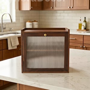 May include: A dark wood cabinet with a glass door featuring vertical lines. The cabinet has a gold-colored knob and is set on a white countertop. The background shows a kitchen with wooden cabinets and a white tile backsplash.