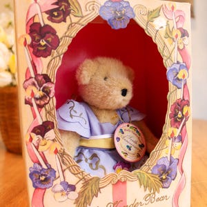 May include: A Muffy VanderBear Tenth Anniversary teddy bear in a decorative box. The bear wears a lavender dress with a yellow sash and a tag. The box is adorned with floral and ribbon designs, with the text "Muffy Vander Bear Tenth Anniversary".
