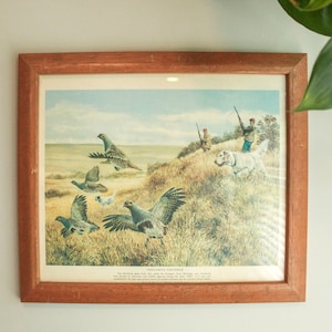 May include: A framed print featuring a landscape scene with multiple birds in flight and on the ground, two hunters with rifles, and a white dog. The artwork is enclosed in a simple wooden frame with a light brown finish and includes text below.