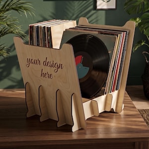 May include: A light-coloured wooden vinyl record storage rack featuring a slotted design for holding multiple records. The front panel displays the text 'your design here' in a dark script font. The rack is constructed from interlocking panels.