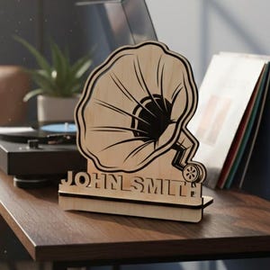 May include: A wooden gramophone-shaped decor piece with the name "JOHN SMITH" engraved on the base. The decor is light brown with black detailing and sits on a dark brown table. A record player and vinyl records are in the background.