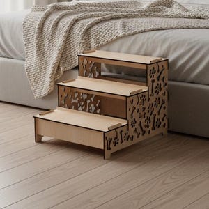 Puede incluir: Un taburete de madera de tres escalones para mascotas con recortes de huellas de patas en el lateral. Los escalones son de color marrón claro y están diseñados para ayudar a perros o gatos pequeños a alcanzar una cama. El taburete está colocado sobre un suelo de madera clara.