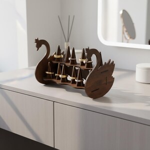 May include: A dark brown wooden ring holder shaped like two swans facing each other. The ring holder has multiple small, angled posts to display rings. Several rings are displayed on the posts. The ring holder sits on a white surface.