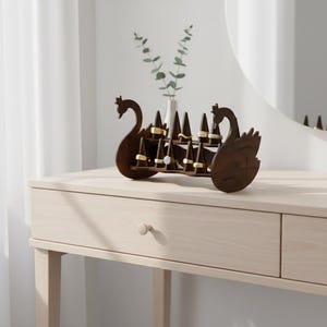 May include: A dark brown wooden ring holder shaped like a swan. The holder has multiple triangular posts to display rings. The swan-shaped holder is on a light-colored wooden table, with a vase of greenery in the background.