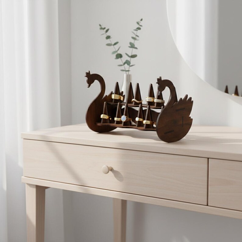 May include: A dark brown wooden ring holder shaped like a swan. The holder has multiple triangular posts to display rings. The swan-shaped holder is on a light-colored wooden table, with a vase of greenery in the background.