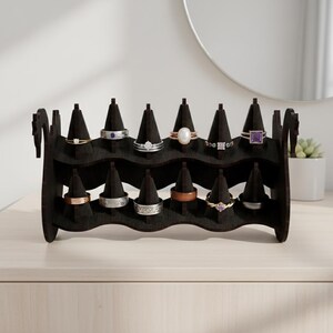 May include: A dark brown wooden ring display stand with multiple ring holders. The stand is holding various rings with different designs and gemstones. The stand is on a light-colored surface.