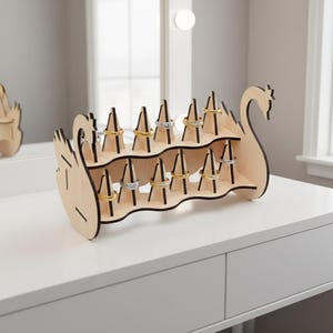 May include: A wooden ring holder in the shape of a swan, with multiple slots to display rings. The holder is made of light-colored wood with black accents and is placed on a white surface. Several rings are displayed on the holder.