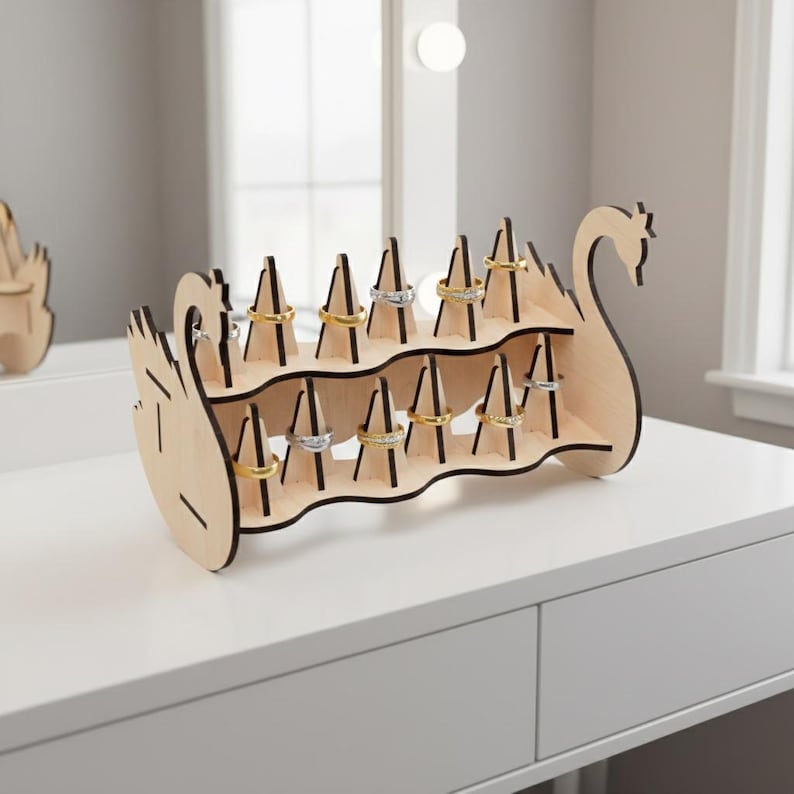 May include: A wooden ring holder in the shape of a swan, with multiple slots to display rings. The holder is made of light-colored wood with black accents and is placed on a white surface. Several rings are displayed on the holder.