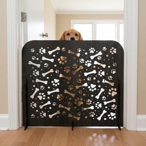 May include: A black wooden pet gate featuring a design of dog bones and paw prints. A golden retriever is visible above the gate. The gate is in a doorway, with a wooden floor. The gate's height is approximately 76 cm.