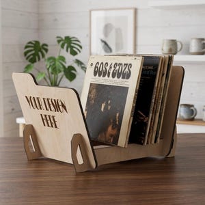 Può includere: Un supporto per dischi in vinile in legno chiaro con base piatta e due pannelli laterali. Il pannello frontale presenta il testo "YOUR DESIGN HERE" inciso al laser con un carattere stilizzato. Il supporto contiene diversi dischi in vinile.