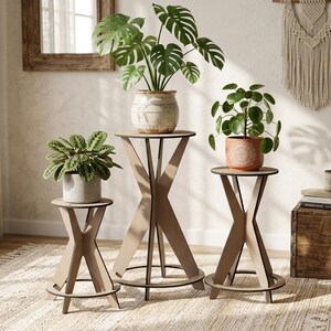 May include: Three light brown wooden plant stands of varying heights, each featuring a circular top, a cross-shaped base, and a circular floor ring. The stands hold a green monstera plant, a green patterned leaf plant, and a green pilea plant.